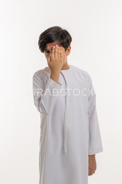 Injury and bruises, receiving medical care, feeling tired and exhausted, being exposed to violence and harm, portrait of an Arab Gulf Emirati boy wearing a kandura, putting his hand on his eye with gestures of severe pain, white background