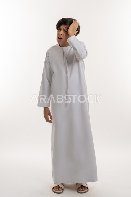 الحاجة إلى تلقي الرعاية الطبية، الشعور بالتعب والارهاق، التعرض للعنف والاذى، بورتريه لفتى عربي خليجي اماراتي يرتدي الكندورة يضع يده على رأسه  بايماءات الالم الشديد، الاصابة بالجروح والكدمات، صورة لكامل الجسم، خلفية بيضاء