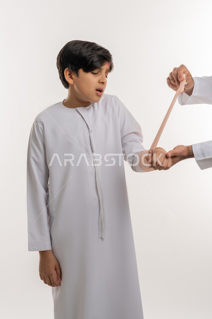 مشاكل صحية وكدمات جسدية، ايماءات الألم والحزن، تلقي الرعاية الطبية المنزلية، بورتريه لفتى عربي خليجي اماراتي يرتدي الكندورة يقوم الده بلف ذراعه برباط مطاطي، الاصابة بالكسور والرضوض، خلفية بيضاء