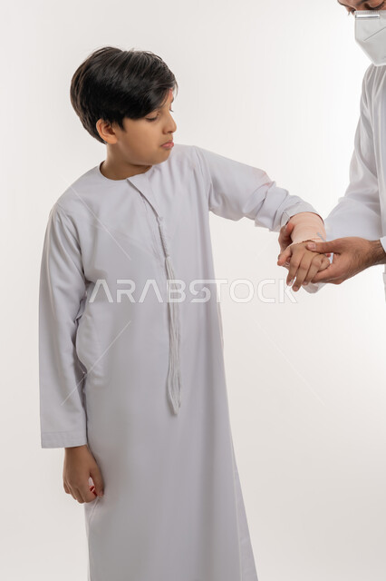 تلقي الرعاية الطبية المنزلية، مشاكل صحية وكدمات جسدية، بورتريه لفتى عربي خليجي اماراتي يرتدي الكندورة يقوم الده بلف ذراعه برباط مطاطي، الاصابة بالكسور والرضوض، تعابير وايماءات الألم والحزن، خلفية بيضاء