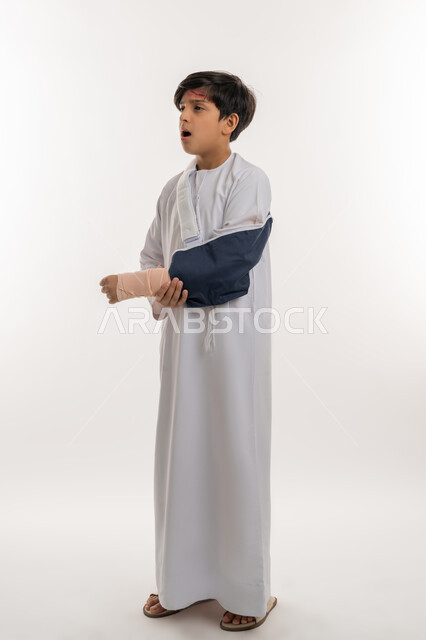 الاصابة بالكسور والرضوض، مشاكل صحية وكدمات جسدية، بورتريه من الجانب لفتى عربي خليجي اماراتي يرتدي الكندورة وحمالة الذراع يمسك يده بتعابير الألم والحزن، التعرض للعنف الأسري، الحاجة الى الرعاية الطبية والعلاج، صورة لكامل الجسم، خلفية بيضاء
