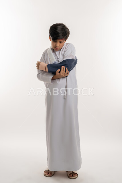 مشاكل صحية وكدمات جسدية، بورتريه لفتى عربي خليجي اماراتي يرتدي الكندورة وحمالة الذراع يمسك يده بتعابير الألم والحزن، التعرض للعنف الأسري، الاصابة بالكسور والرضوض، الحاجة الى الرعاية الطبية والعلاج، صورة لكامل الجسم، خلفية بيضاء