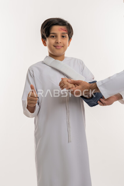 تعابير الاعجاب والرضى، الاصابة بالكسور والرضوض، مشاكل صحية وكدمات جسدية، بورتريه لفتى عربي خليجي اماراتي يرتدي الكندورة وحمالة الذراع يقوم بتلقي الرعاية الطبية المنزلية يرفع اصبع الابهام بايماءات الشعور بالتحسن، خلفية بيضاء