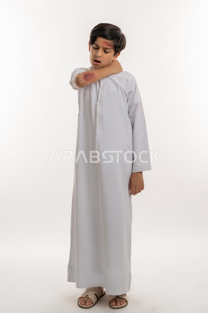 التعرض للعنف والاذى، الشعور بالالم الشديد، الحاجة الى الرعاية الطبية والعلاج، اصابات وكدمات جسدية، بورتريه لصبي عربي خليجي اماراتي مصاب يرتدي الكندورة يقوم بالنظر الى الجروح بتعابير التعب والارهاق، صورة لكامل الجسم، خلفية بيضاء
