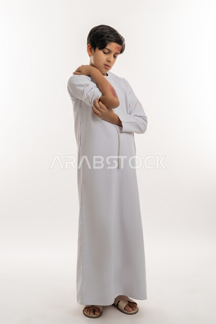 التعرض للعنف والاذى، الحاجة الى الرعاية الطبية والعلاج، الشعور بالالم الشديد، اصابات وكدمات جسدية، بورتريه لصبي عربي خليجي اماراتي مصاب يرتدي الكندورة يقوم بالنظر الى الجروح بتعابير التعب والارهاق، صورة لكامل الجسم، خلفية بيضاء