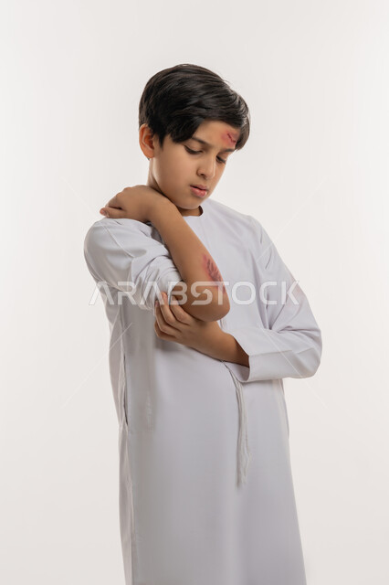 الحاجة الى الرعاية الطبية والعلاج، الشعور بالالم الشديد، التعرض للعنف والاذى، اصابات وكدمات جسدية، بورتريه لصبي عربي خليجي اماراتي مصاب يرتدي الكندورة يقوم بالنظر الى الجروح بتعابير التعب والارهاق، خلفية بيضاء