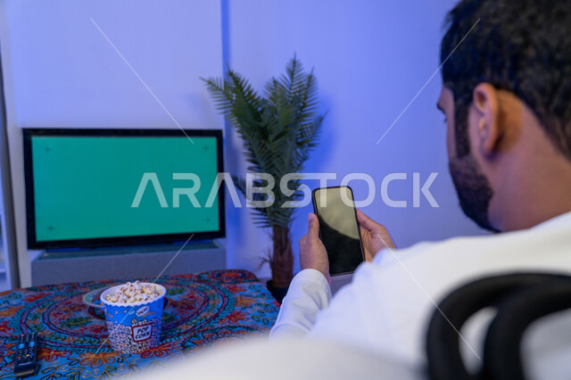 استخدام تقنية السمارت على الهاتف للتحكم بالتلفاز، اختيار فلم من الهاتف لاستعراضه على شاشة التلفاز, قضاء وقت ممتع في المنزل, صورة من الخلف لرجل عربي اماراتي خليجي يرتدي الثوب الأبيض التقليدي يحمل بيده الهاتف المحمول وامامه التلفاز بشاشة خضراء فارغة، كروما