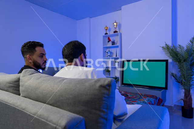 مفهوم التسليه والترفيه الجماعي، قضاء الوقت في المنزل مع الأصدقاء، صورة من الخلف لرجلان عربيان خليجيان اماراتيان يجلسان على الأريكة في غرفة المعيشة يستمتعان بمشاهدة الفيلم على التلفاز بخلفية كروما خضراء