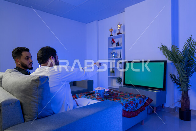قضاء الوقت في المنزل مع الأصدقاء، صورة من الخلف لرجلان عربيان خليجيان اماراتيان يجلسان على الأريكة في غرفة المعيشة يستمتعان بمشاهدة الفيلم على التلفاز بخلفية كروما خضراء، مفهوم التسليه والترفيه الجماعي