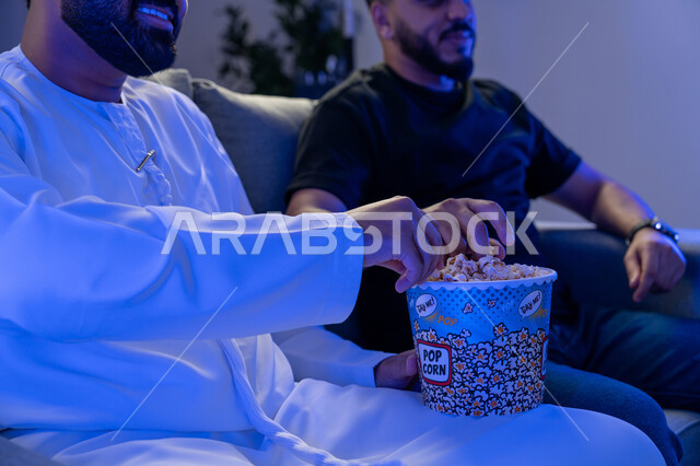 Group home entertainment activity, two young Arab Emirati Gulf men enjoying watching a movie on TV and eating popcorn, immersion and interaction with the movie, having a good time with friends, enjoying eating popcorn