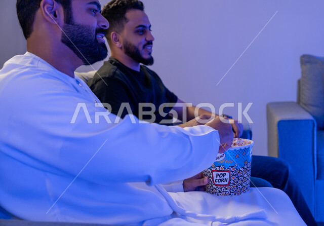 قضاء وقت ممتع في المنزل، تناول الفوشار والتسالي اثناء متابعة الأفلام والمسلسلات، الاستمتاع بالأنشطة الترفيهيه في عطلة نهاية الاسبوع، رجلان عربيان خليجيان اماراتيان يستمتعان بتناول البوب كورن ومشاهدة التلفاز
