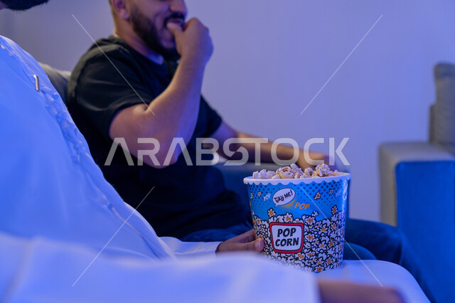 الاستمتاع بالأنشطة الترفيهيه في عطلة نهاية الاسبوع، قضاء وقت ممتع في المنزل، تناول الفوشار والتسالي اثناء متابعة الأفلام والمسلسلات، رجلان عربيان خليجيان اماراتيان يستمتعان بتناول البوب كورن ومشاهدة التلفاز
