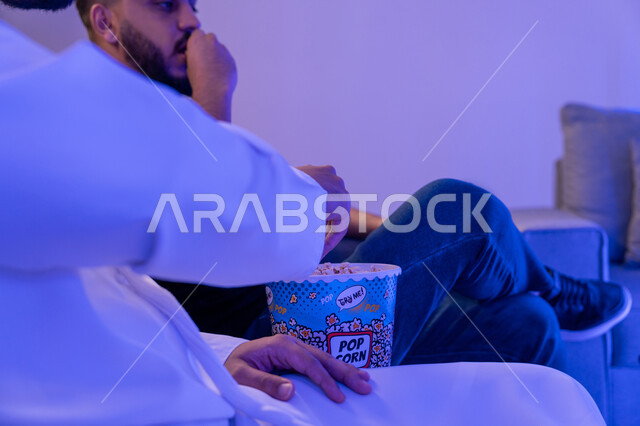 الاستمتاع بتناول الفشار أثناء مشاهدة الفلم , انشطة عطلة نهاية الأسبوع , صديقان عربيان سعوديان خليجيان يستمتعان بوقت الفراغ , إيماءات وجه تدل على السعادة والاندماج بمشاهدة الأفلام والمسلسلات التلفزيونية, قضاء وقت ممتع في المنزل