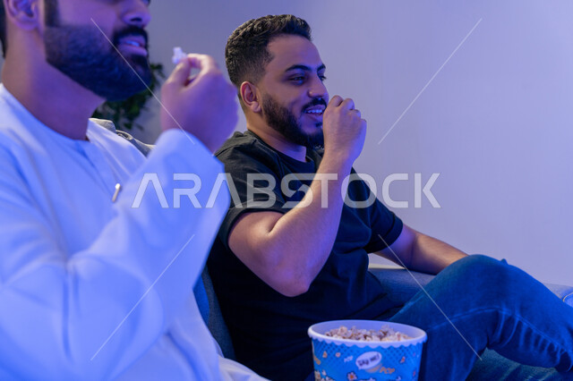 قضاء وقت ممتع في المنزل، مفهوم التسليه والترفيه الجماعي, تصوير مقرب لصديقان عربيان خليجيان امارتيان يجلسان على أريكه مريحه في غرفه المعيشه امام شاشه التلفاز يتناولان البوب كورن، تعابير تدل على السعاده والسرور