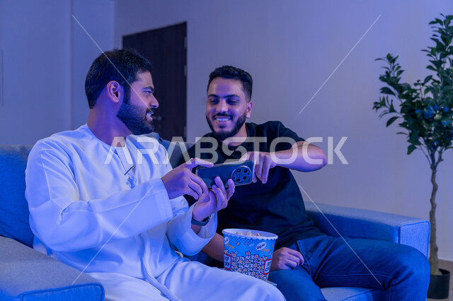 قضاء وقت ممتع مع الأصدقاء, رجلان عربيان امارتيان خليجيان يستمتعان بمشاهدة فلم على الهاتف المحمول، نقاش حول مشهد في الهاتف, نشاط منزلي ترفيهي جماعي, الإستمتاع بأكل الفشار