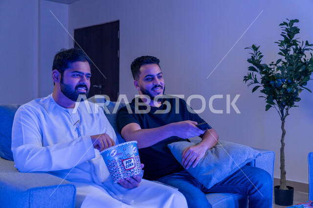 إيماءات وجه تدل على الإندماج والتفاعل مع الفلم, صديقان عربيان امارتيان خليجيان يستمتعان بمشاهدة فلم على جهاز التلفاز، نشاط منزلي ترفيهي جماعي, قضاء وقت ممتع مع الأصدقاء, الإستمتاع بأكل الفشار