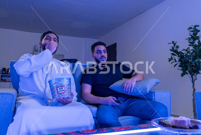 نشاط منزلي ترفيهي جماعي, شابان عربيان امارتيان خليجيان يستمتعان بمشاهدة فلم على جهاز التلفاز، مشاهدة فلم رعب, ايماءات وجه تدل على الخوف والدهشه, قضاء وقت ممتع مع الأصدقاء, الإستمتاع بأكل الفشار