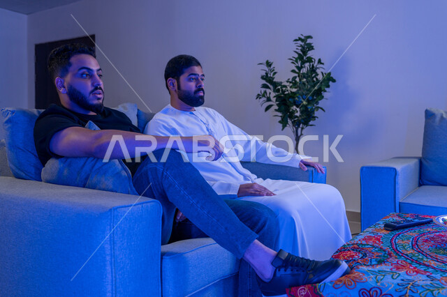 قضاء وقت ممتع مع الأصدقاء اثناء مشاهده التلفاز، مفهوم التسليه والترفيه الجماعي, صديقان عربيان خليجان اماراتيان يجلسان على أريكه مريحه في غرفه المعيشه امام شاشه التلفاز ينظرون بتعابير تدل على الاندماج والتركيز