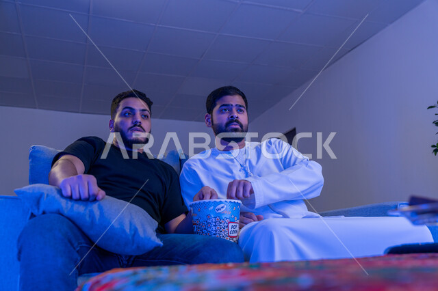 مفهوم التسليه والترفيه الجماعي, قضاء وقت ممتع بتناول الفشار اثناء مشاهده التلفاز، صديقان عربيان خليجان اماراتيان يجلسان على أريكه مريحه في غرفه المعيشه امام شاشه التلفاز يحملان بيدهما كوب مليئ بالبوب كورن، تعابير تدل على الاندماج والتركيز