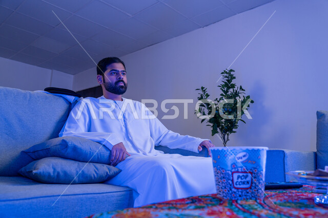 مفهوم التسليه والترفيه، ايماءات وتعابير الاندماج والتركيز، قضاء أوقات ممتعة ومسلية في المنزل، الاستمتاع بمشاهدة الأفلام والمسلسلات، رجل عربي خليجي اماراتي يرتدي الزي التقليدي يجلس على اريكة مريحة يقوم بمشاهدة التلفاز وأمامه كوب مليئ بالفشار