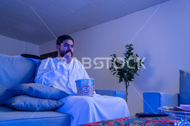 ايماءات وتعابير الاندماج والتركيز، قضاء أوقات ممتعة ومسلية في المنزل، الاستمتاع بمشاهدة الأفلام والمسلسلات، رجل عربي خليجي اماراتي يرتدي الزي التقليدي يجلس على اريكة مريحة يقوم بمشاهدة التلفاز وتناول الفوشار