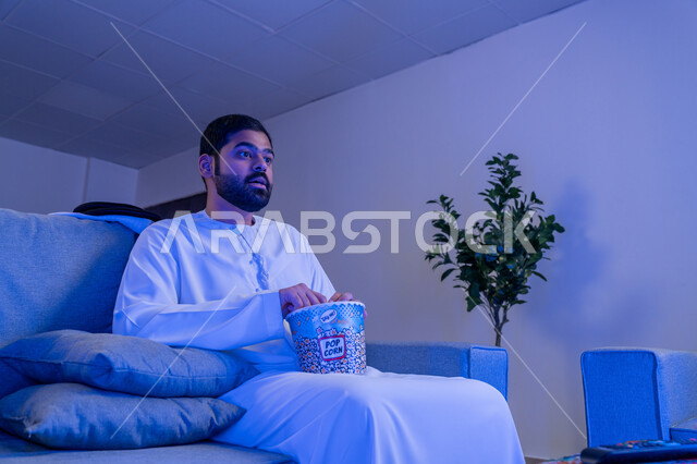 قضاء أوقات ممتعة ومسلية في المنزل، الاستمتاع بمشاهدة الأفلام والمسلسلات، رجل عربي خليجي اماراتي يرتدي الزي التقليدي يجلس على اريكة مريحة يقوم بمشاهدة التلفاز وتناول الفوشار، ايماءات وتعابير الاندماج والتركيز