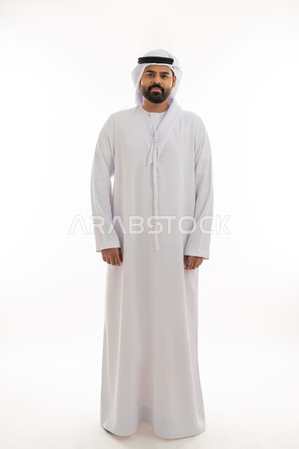 مفهوم الاناقه والاهتمام بالمظهر الخارجي، الوقوف باستقامه والنظر الى الكاميرا بتعابير الثقه بالنفس، بورتريه لرجل عربي خليجي اماراتي يرتدي الكندوره والغتره البيضاء الاماراتيه يقف بايماءات الرجولة، الزي الإماراتي الشعبي، الجسم بالطول الكامل، خلفية بيضاء