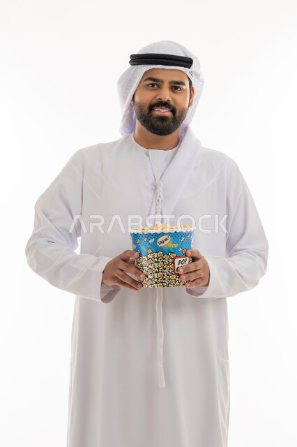 مفهوم التسليه والترفيه، قضاء أوقات ممتعة في عطلة نهاية الاسبوع، أنشطة وفعاليات مسلية، الاستعداد لمشاهدة فيلم في السينما، بورتريه لشاب عربي خليجي اماراتي يرتدي الكندورة والغترة يحمل بيده علبة من الفشار المقرمش ينظر إلى الكاميرا بايماءات السرور، خلفية بيضاء
