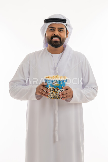 Having fun at home, home entertainment activities, eating snacks, portrait of an Emirati Gulf Arab man wearing an Emirati kandura and ghutra holding a box of crunchy popcorn, enjoying watching movies and series, following TV shows, white background