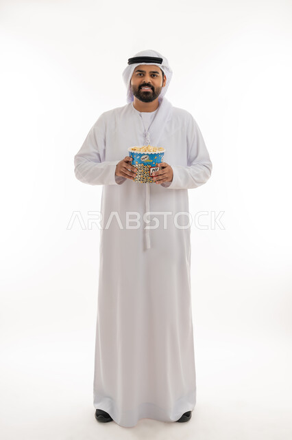 انشطة ترفيهية منزلية، تناول الوجبات الخفيفة والسناك، بورتريه لرجل عربي خليجي اماراتي يرتدي الكندورة الاماراتية والغترة يحمل بيده علبة من الفوشار المقرمش، الاستمتاع بمشاهدة الأفلام والمسلسلات، متابعة برامج التلفاز، الجسم بالطول الكامل، خلفية بيضاء