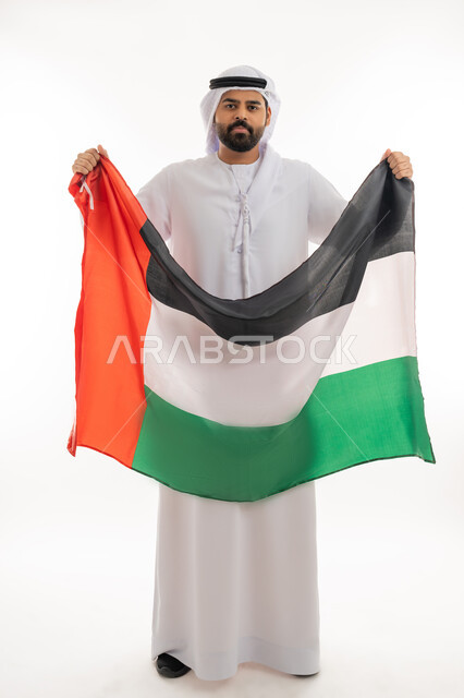 هوية اليوم الوطني الاماراتي 51، الإحتفال باليوم الوطني الإماراتي 2 ديسمبر، بورتريه لرجل عربي خليجي إماراتي يرتدي الكندورة والغترة يحمل علم الإمارات العربية المتحدة وينظر الى الكاميرا بايماءات الاعتزاز والفخر بإنجازات البلاد، الجسم بلطول الكامل، خلفية بيضاء