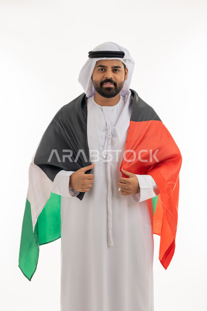 Commemorating the Independence Day of the Emirates, December 2, looking at the camera with gestures of pride and confidence, the identity of the 51st UAE National Day, a portrait of an Arab Gulf Emirati man wearing a kandura and a ghutra carrying the UAE flag on his back, love and belonging to the homeland, white background