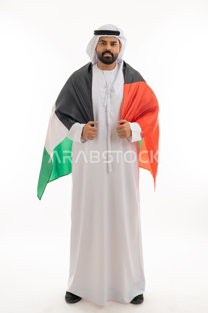 النظر الى الكاميرا بايماءات الاعتزاز والثقة، هوية اليوم الوطني الاماراتي 51، احياء ذكرى استقلال الإمارات 2 ديسمبر، بورتريه لرجل عربي خليجي إماراتي يرتدي الكندورة والغترة يقوم بحمل علم الإمارات على ظهره، الحب والانتماء للوطن، الجسم بالطول الكامل، خلفية بيضاء