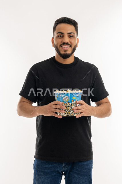 الاستعداد لمشاهدة فيلم في السينما، قضاء أوقات ممتعة في عطلة نهاية الاسبوع، أنشطة وفعاليات مسلية، تعابير السرور والفرح، بورتريه لشاب عربي خليجي اماراتي يحمل بيده علبة من الفشار المقرمش ينظر إلى الكاميرا بايماءات السعادة، خلفية بيضاء