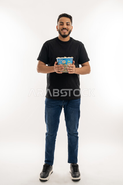 قضاء أوقات ممتعة في عطلة نهاية الاسبوع، أنشطة وفعاليات مسلية، الاستعداد لمشاهدة فيلم في السينما، تعابير السرور والفرح، بورتريه لشاب عربي خليجي اماراتي يحمل بيده علبة من الفشار المقرمش ينظر إلى الكاميرا بايماءات السعادة، الجسم بالطول الكامل، خلفية بيضاء