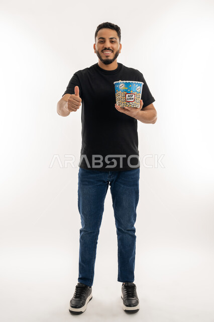 أنشطة وفعاليات مسلية، الاستعداد لمشاهدة فيلم في السينما، تعابير السرور والفرح، قضاء أوقات ممتعة في عطلة نهاية الاسبوع، بورتريه لشاب عربي خليجي اماراتي يحمل بيده علبة من الفشار المقرمش يرفع ابهامه بإيماءات الرضا والاعجاب، الجسم بالطول الكامل، خلفية بيضاء