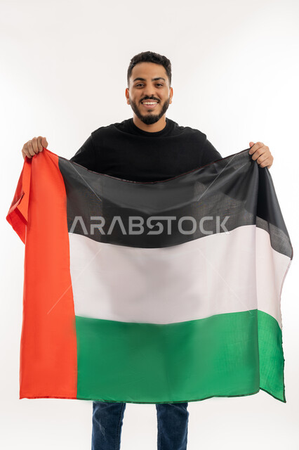 الإحتفال بالعيد الوطني الإماراتي 2 ديسمبر، إحياء ذكرى الإستقلال لدولة الإمارات العربية المتحدة، حب الوطن والعلم، بورتريه لرجل عربي خليجي إماراتي يرتدي زي كاجوال يحمل علم الإمارات ينظر إلى الكاميرا بايماءات الفخر والاعتزاز، خلفية بيضاء