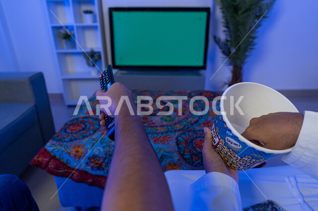 نشاط منزلي ترفيهي جماعي, قضاء وقت ممتع مع الأصدقاء، صورة من الخلف لأيدي شابان عربيان اماراتيان خليجيان يشاهدان شاشة تلفاز خضراء فارغة يستمتعان بأكل الفشار، الاهتمام بمتابعة الأفلام والمسلسلات، كروما
