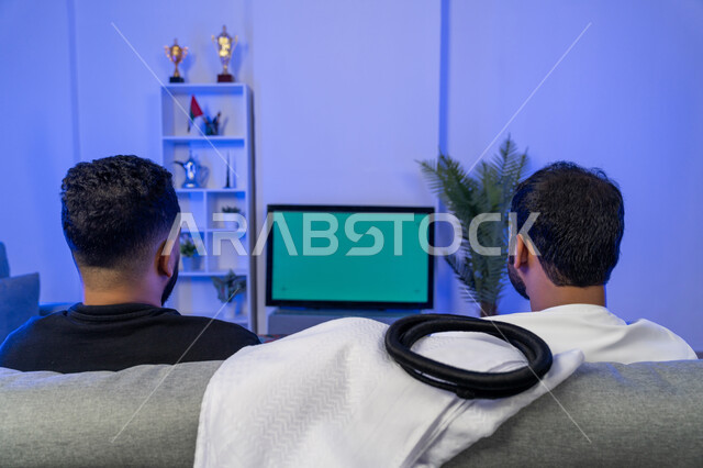 مفهوم التسليه والترفيه الجماعي، قضاء الوقت في المنزل مع الأصدقاء، صورة من الخلف لرجلين عربيين خليجيين اماراتيين يجلسان على الأريكة في غرفة المعيشة يستمتعان بمشاهدة الافلام على التلفاز بخلفية خضراء، الاستمتاع بالأنشطة الترفيهيه في عطلة نهاية الاسبوع، كروما