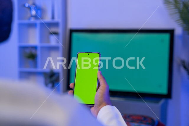 قضاء أوقات ونشاطات ممتعة في المنزل، استخدام تقنية السمارت على الهاتف للتحكم بالتلفاز, اختيار فلم من الهاتف لاستعراضه على شاشة التلفاز, صورة مقربة من الخلف لشاب عربي اماراتي خليجي يحمل بيده الهاتف المحمول بشاشة خضراء فارغة، كروما