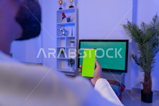 استخدام تقنية السمارت على الهاتف للتحكم بالتلفاز, اختيار فلم من الهاتف لاستعراضه على شاشة التلفاز, صورة مقربة من الخلف لشاب عربي اماراتي خليجي يحمل الهاتف المحمول بشاشة خضراء فارغة، قضاء وقت ممتع في المنزل، كروما