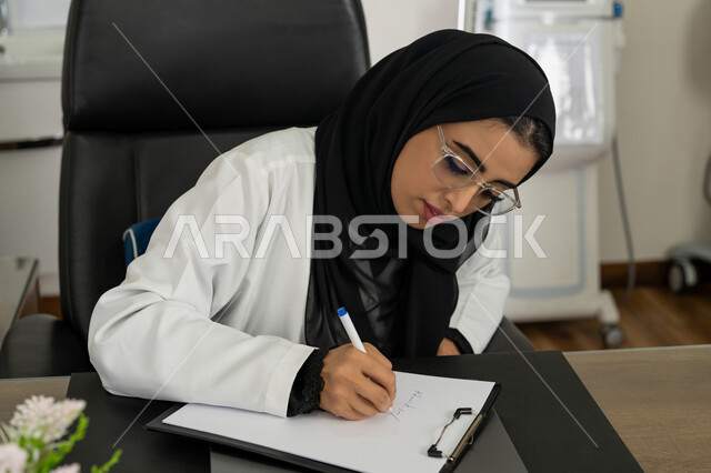 الاطلاع على نتائج التحاليل, تقديم الاستشارات في المراكز الطبيه في الامارات العربية المتحدة, متابعة السجل الطبي للمراجعين, العمل في القطاعات الصحية, صورة مقربة لطبيبة عربية خليجية اماراتية محجبة ترتدي المعطف الطبي تقوم بكتابة وصفة للمريض, خلفية بيج