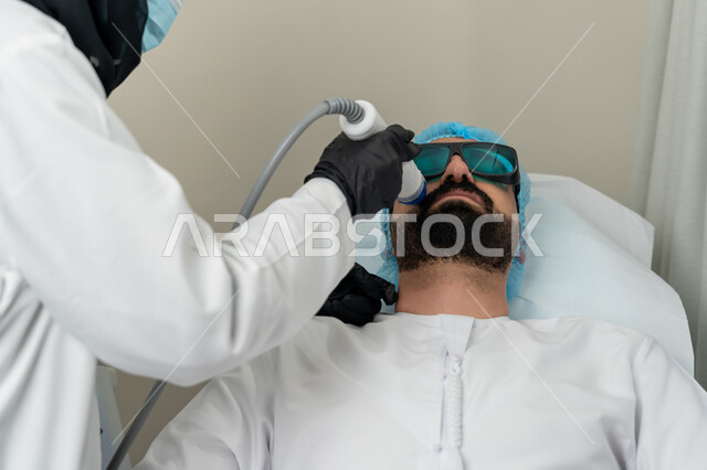 جلسة ليزر في عيادة خاصة، سيد عربي اماراتي خليجي يضع النظارة للحماية في موعد بعيادة طبيبة مختصة بالتنظيف والعناية بالبشرة، طبيبة عربية خليجية اماراتية محجبة تستخدام احدث التقنيات لعلاج الوجه، تقديم الخدمات والرعاية الصحية, خلفية بيضاء