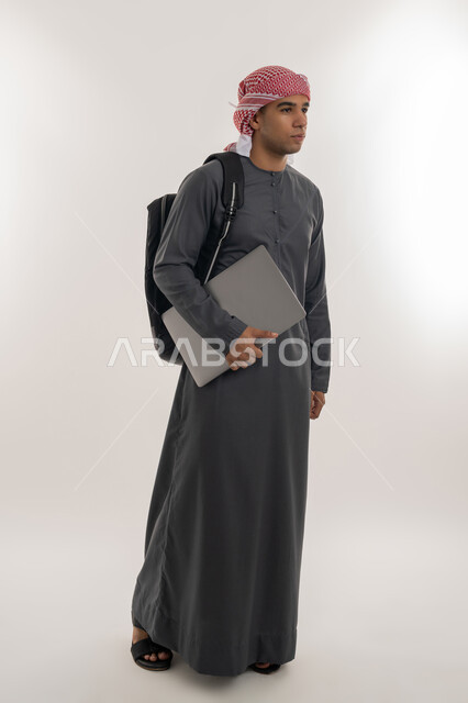 دمج التقنيات الحديثة بالتعليم الأكاديمي, المناهج والمواد التعليمية, الجامعات و الكليات في الإمارات, بورتريه لطالب جامعي عربي خليجي إماراتي يرتدي الكندورة والعمامة يحمل حقيبة على ظهره وبيده جهاز الحاسوب المحمول, الجسم بالطول الكامل, خلفية رمادية
