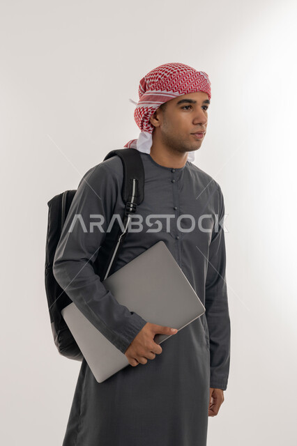 دمج التقنيات الحديثة بالتعليم الأكاديمي, المناهج والمواد التعليمية, الجامعات و الكليات في الإمارات, بورتريه لطالب جامعي عربي خليجي إماراتي يرتدي الكندورة والعمامة يحمل حقيبة على ظهره وبيده جهاز الحاسوب المحمول, خلفية رمادية