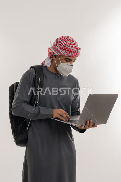 التعليم عن بعد للحفاظ على السلامة, القيام بالإجراءات الوقائية لتجنب الإصابة بفايروس كورونا, استخدام الأجهزة التقنية الحديثة، بورتريه لطالب جامعي عربي خليجي إماراتي يرتدي الكندورة و العمامة يضع الكمامة ويقوم باستعمال الحاسوب, خلفية رمادية