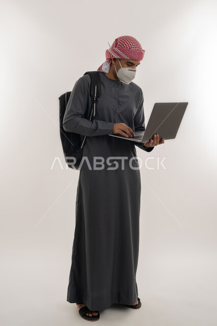 التعليم عن بعد للحفاظ على السلامة, القيام بالإجراءات الوقائية لتجنب الإصابة بفايروس كورونا, استخدام الأجهزة التقنية الحديثة، بورتريه لطالب جامعي عربي خليجي إماراتي يرتدي الكندورة و العمامة يضع الكمامة ويقوم باستعمال الحاسوب, الجسم بالطول الكامل, خلفية رمادية