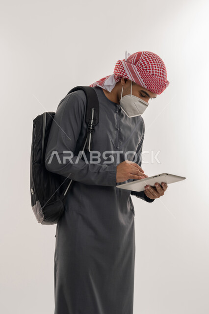 القيام بالإجراءات الوقائية لتجنب الإصابة بفايروس كورونا, التعليم عن بعد للحفاظ على السلامة، الكليات الاكاديمية الإماراتية، بورتريه لطالب جامعي عربي خليجي إماراتي يرتدي الكندورة و العمامة يضع الكمامة يحمل بيده الجهاز اللوحي, خلفية رمادية