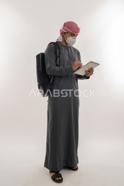 التعليم عن بعد للحفاظ على السلامة, القيام بالإجراءات الوقائية لتجنب الإصابة بفايروس كورونا، الجامعات الاكاديمية الإماراتية، بورتريه لطالب جامعي عربي خليجي إماراتي يرتدي الكندورة و العمامة يضع الكمامة يحمل بيده الجهاز اللوحي, الجسم بالطول الكامل, خلفية رمادية