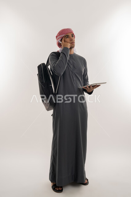 Planning and thinking about the future, finding solutions to problems, using modern technical devices, an Arab Gulf Emirati university student wearing a kandura and turban, carrying a bag on his back, holding a tablet and placing his hand on his head with expressions of contemplation and concentration, full body, gray background